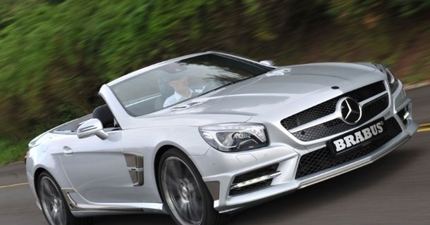 Hvid Mercedes-Benz SL-model cabriolet uden tag, fotograferet under kørsel på landsvej med grønt område i baggrunden
