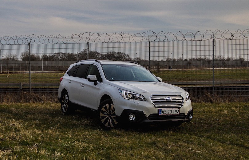 Hvid stationcar-SUV parkeret på grønt område foran høj metalindhegning med pigtråd