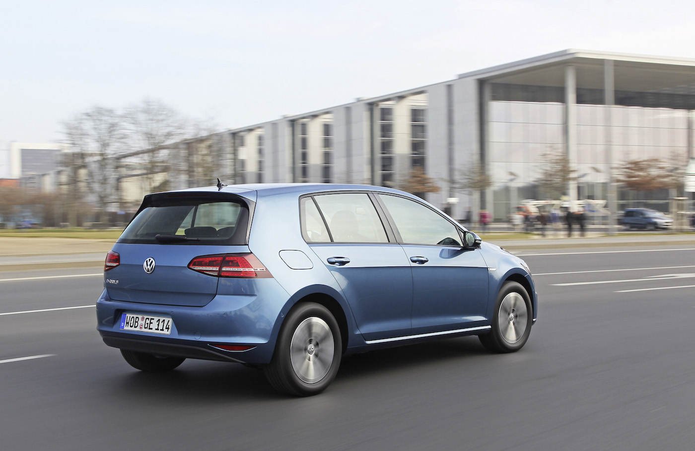 Blå Volkswagen Golf ses fra siden mens den kører på en bred vej foran en moderne bygning