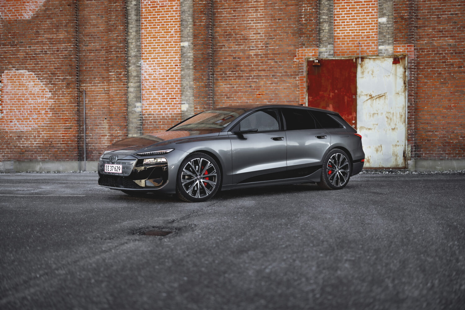 En grå Audi stationcar holder parkeret foran en rød murstensvæg med en rusten metaldør i baggrunden. Bilens skarpe linjer og sporty fælge fremhæver designet i en rå urban setting der passer til Bilsektionen og Audi.