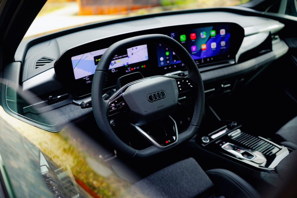 Interiør af en Audi med fokus på rattet med Audi logo og et digitalt instrumentpanel med navigation og apps på skærmen. Den moderne kabine fremhæver teknologi og komfort i en kontekst der passer til Bilsektionen og Audi.