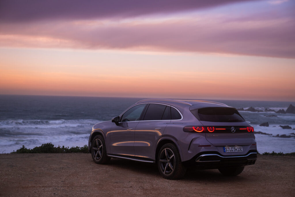 En sølvgrå Mercedes SUV holder parkeret ved kysten med udsigt over havet ved solnedgang og baglygterne tændt. Den rolige scene fremhæver bilens elegante design i en eksklusiv setting der passer til Bilsektionen og Mercedes.