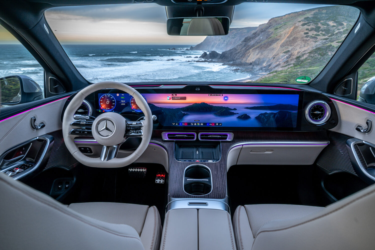 Interiør af en Mercedes med lyst læder, ambient belysning og et bredt digitalt display der viser navigation og landskab. Udsigten gennem forruden mod kyst og klipper understreger luksus og teknologi i en kontekst der passer til Bilsektionen og Mercedes.