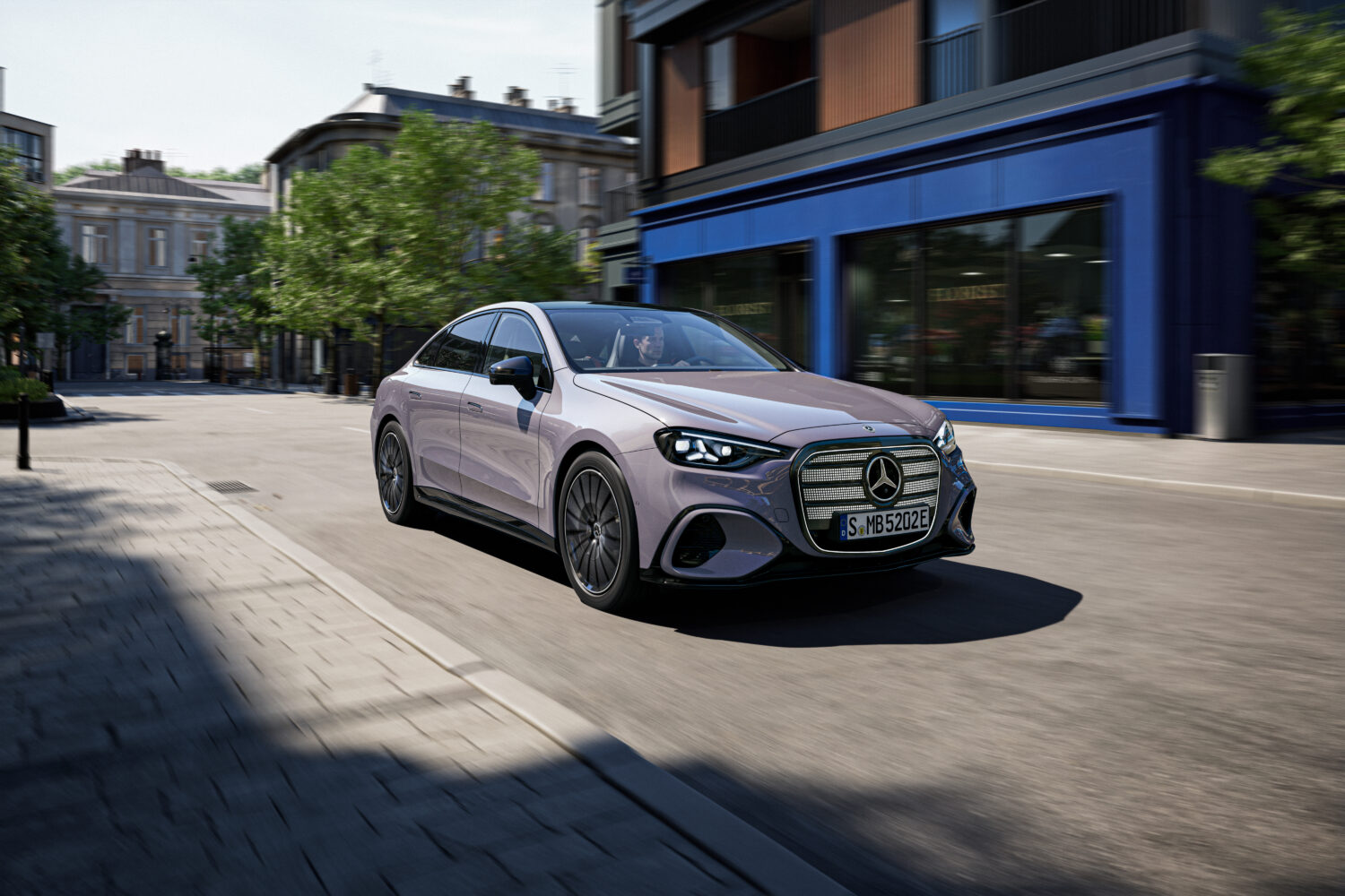 Interiør af en Mercedes med sort kabine, blå ambient belysning og et bredt digitalt display der viser navigation og infotainment. Udsigten gennem ruden til en bygade med butiksvinduer giver kontekst til en moderne køreoplevelse der passer til Bilsektionen og Mercedes.