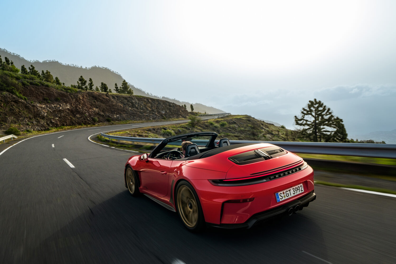 En rød Porsche cabriolet kører gennem et sving på en bjergvej med udsigt over grønne skråninger og tågede bjerge i baggrunden. Bilen ses bagfra med føreren i fokus og fremhæver fart og køreglæde i en scene der passer til Bilsektionen og Porsche.